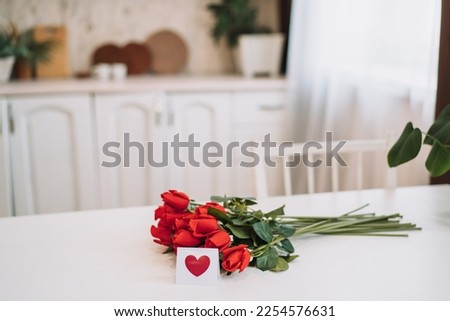 Bouquet of red roses and small card with painted red heart on white table.