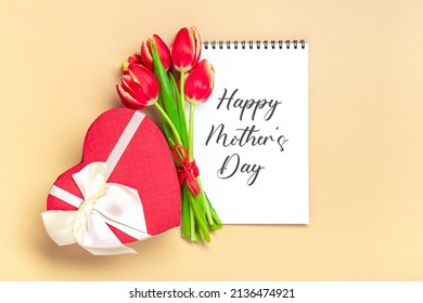 Bouquet of red tulips, gift box heart shaped, text Happy Mother's Day on open  white notepad on beige background Top view Flat lay Holiday greeting card