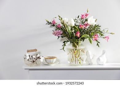 Bouquet de tulipes blanches, eustome rose, hyacinthe, eucalyptus dans un vase de verre déversé sur le panneau d'une cheminée artificielle. Thé dans une tasse, figurines de fidèles d'un oiseau et d'un ange. Espace vide.