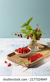 Bouquet of wild strawberry berries with leaves  in glass vase and lot of berries on wooden tray. Summer still life with wild strawberries on  blue background. Copy space. 