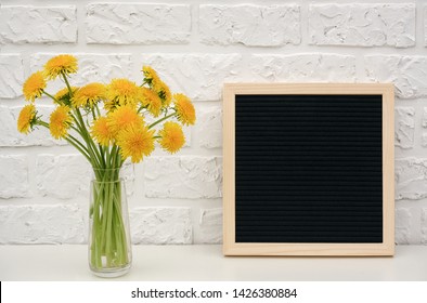 Bouquet yellow dandelion in vase and blank black letter board on table against white brick wall. Mockup template for your text, design. Concept Women's day, Mothers Day, Hello summer , Hello spring.