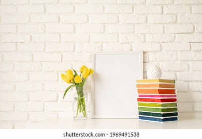 Bouquet of yellow tulips in a glass vase, pile of colorful books and blank photo frame on a white brick wall background. Mock up design - Shutterstock ID 1969491538