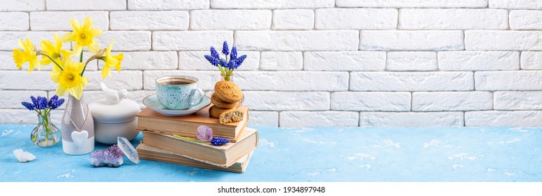 Bouquets Of Spring Flowers, Coffee Cup, Books, Cookies, Gemstones Crystal Minerals On Blue Table On White Brick Background. Reading And Breakfast. Concept Spring, Gems, Hygge And Cozy Home Interior
