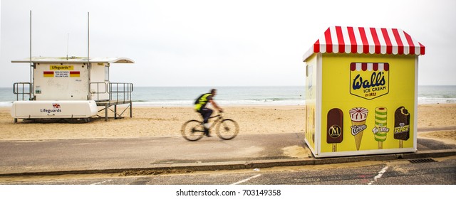 BOURNEMOUTH, UK - AUGUST 22, 2017: Cyclist Rides Passed A Life Guard Station Towards A Walls Ice Cream Stand On Bournemouth Beach