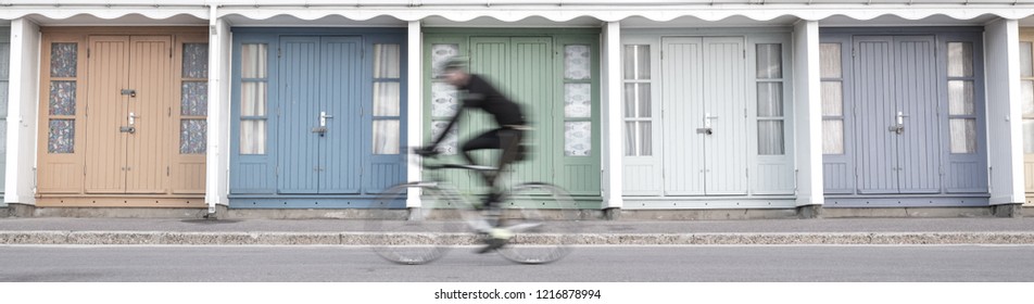 Bournemouth UK, October 2018. People Cycling In Front Of Colourful Beach Huts Located On The Promenade On The Bournemouth Sea Front. Cyclists Are Out Of Focus. 