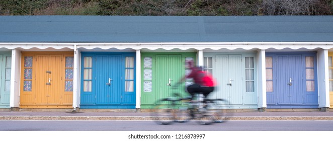 Bournemouth UK, October 2018. People Cycling In Front Of Colourful Beach Huts Located On The Promenade On The Bournemouth Sea Front. Cyclists Are Out Of Focus. 