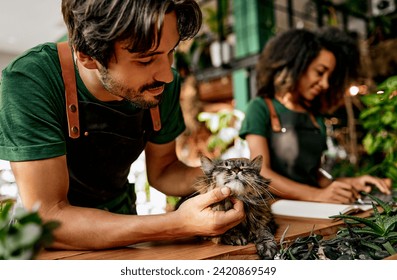 Boutique business. Caucasian bearded man petting fluffy grey cat while african american woman using laptop behind. Friendly married couple enjoying working process at cozy own flower store. - Powered by Shutterstock