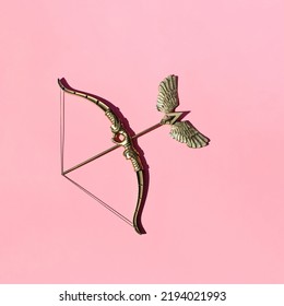 Bow and arrow arranged with golden wings, creative arrangement on pastel pink background. Flying, ambition, passion ideas. 