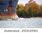 Bow of a lake freighter pushing water down the river with colorful autumn trees in the background along the shoreline