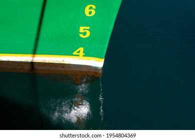 The bow of a large green ship with brown and white stripes at the waterline. The numbers 4, 5, and 6 are painted on the boat in yellow paint as a measurement of meters of water depth or load line.