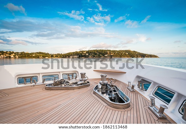 Bow of a new and modern Superyacht with teak deck,\
stainless steel capstans and cleats, with a nice view over blue sky\
and sea