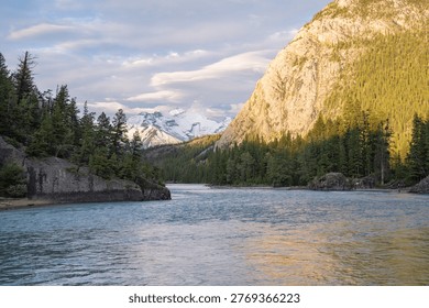 The Bow River flows gently between tall mountains covered with trees in Banff Alberta Canada. The sky shows clouds with some sunlight filtering through. This scene captures nature in a national park.
