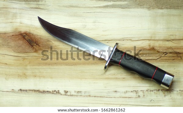 Bowie hunting knife\
on a wooden background. Combat army knife made of high- quality\
stainless steel. Weapons of attack and defense. The polished blade\
of the knife glitters.