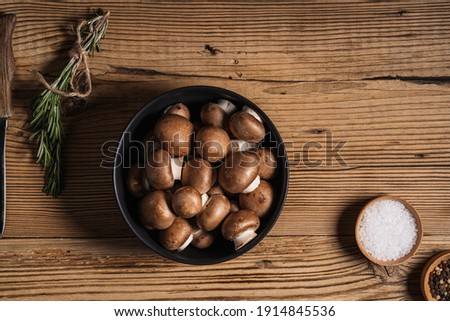 Bowl of baby bella mushrooms and bunch of fresh rosemary on rustic wooden background viewed from above, plant based food