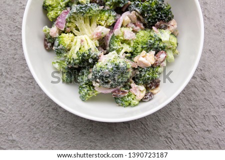 Bowl of broccoli crunch salad with red onions, dried cranberries, cashews and bacon against grey concrete background.