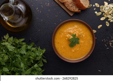 Bowl of carrot and pumpkin soup, piece of bread and green on dark background. Top view