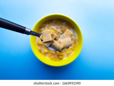 A Bowl Of Cereal In Milk. Cereal Is Made Of Square Pieces, Containing A High Amount Of Sugar. The Bowl Is Yellow. Cereal Is On It Own Against A Blue Background.