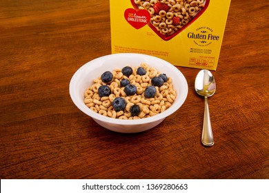 A Bowl Of Cheerios With Blueberries On A Wood Table With Spoon And Box In Background