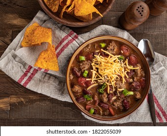 Bowl Of Chili Con Carne With Shredded Cheddar Cheese And Nacho Chips On A Rustic Wooden Table.  Overhead View