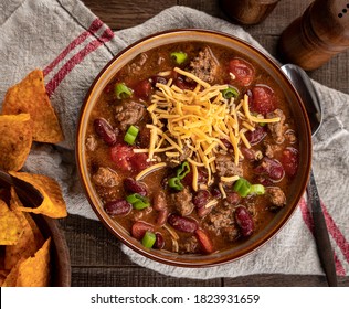 Bowl Of Chili Con Carne With Shredded Ceddar Cheese And Nacho Chips On A Rustic Wooden Table.  Overhead View