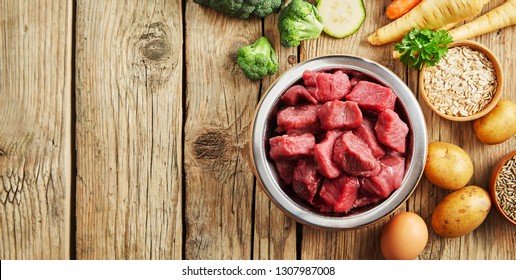 Bowl Of Chopped Raw Meat For A Dog Or Cat On A Rustic Wood Floor Surrounded By Fresh Vegetables For A Healthy Animal Diet