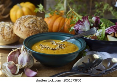 Bowl Of Creamy, Hearty, Savory Sweet Potato Soup, With A Little Ancho Chili Powder; And A Swirl Of Course Ground Spices On Top.  Broken Garlic To Side With Buns And Tossed Salad In Background. 