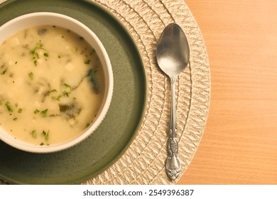 A bowl of creamy soup garnished with herbs, placed on a green plate with a silver spoon beside it on a woven placemat. - Powered by Shutterstock