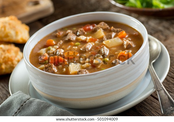 A bowl of delicious beef and\
barley soup with carrots, tomato, potato, celery, and\
peas.
