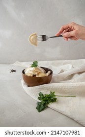 A Bowl Of Dumplings With Cottage Cheese With Sour Cream And Parsley. In Hand Is A Fork With A Dumpling With Cottage Cheese On A Light Gray Background.