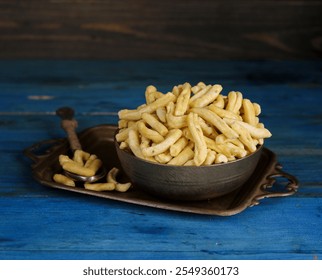 A bowl filled with golden, crispy snacks sits on a rustic tray with a spoon beside it, all placed on a blue wooden surface.