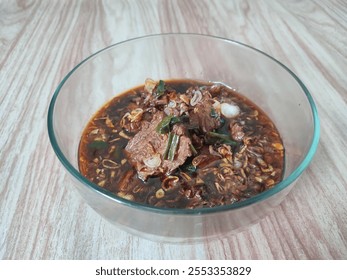 A bowl of flavorful Indonesian beef soup (rawon) with rich black broth, tender meat, and garnished with green onions and fried shallots, served on a rustic table. - Powered by Shutterstock