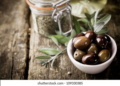 A bowl of fresh black olives and container of dried herbs stand on an old wooden kitchen table for use as ingredients in cooking - Powered by Shutterstock