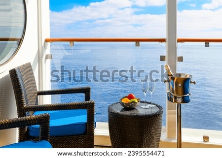 A bowl of fresh fruit, a bottle of champagne in an ice bucket, and two champagne glasses on an oceanfront balcony veranda on a sunny cruise ship. 