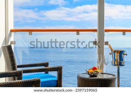 A bowl of fresh fruit, a bottle of champagne in an ice bucket, and two champagne glasses on an oceanfront balcony veranda on a sunny cruise ship. 