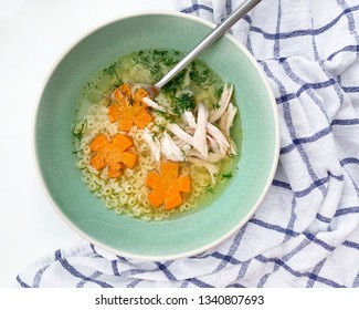 A Bowl Of Fresh Organic Chicken Soup With Small Round Pasta And Flower Shaped Carrots. A Spoon In The Bowl, Ready To Eat. Top View, Horizontal Close Up. Kitchen White Linen Towel On A Side. Kids Meal.