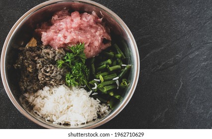 A bowl with fresh and raw dog food made with chicken meat and rumen served with cooked rice and green beans isolated on dark table background with copy space