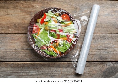 Bowl Of Fresh Salad With Plastic Food Wrap On Wooden Table, Flat Lay