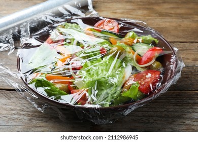 Bowl Of Fresh Salad With Plastic Food Wrap On Wooden Table, Closeup