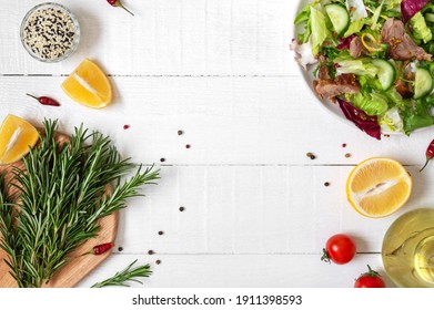 Bowl Of Fresh Salad, Rosemary, Lemon, Spices And Oil On White Wooden Table. Food Background. Ingredients For Cooking With Copy Space. Healthy Eating Concept.