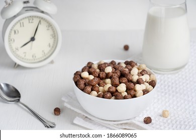 Bowl full of ready-to-eat cold cereals in shape of cocoa balls served with spoon and glass of milk prepared for breakfast on white wooden table with alarm clock in the morning at kitchen. Horizontal