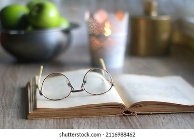 Bowl of green apples, open book, reading glasses, lit candle and flowers on the table. Selective focus. - Powered by Shutterstock