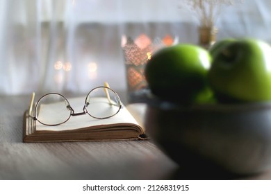 Bowl of green apples, open book, reading glasses, lit candle and flowers on the table. Selective focus. - Powered by Shutterstock