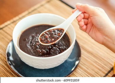 A Bowl Of Homemade Red Bean Soup And Purple Rice Porridge With A Spoon Held By A Female's Hand. 
