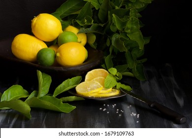 A bowl of lemons and limes with a small plate of sliced lemons in the foreground.  Lemons and limes with leaves against a black background. black handled knife rests on the plate.