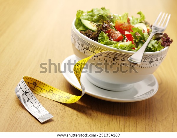 A bowl of mixed salad with a tape\
measure for healthy eating on a wooden\
background.
