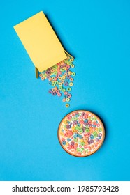 Bowl With Multicolored Cereals And Milk On A Blue Table, Above View. Cereal Box Overturned On A Colored Table.