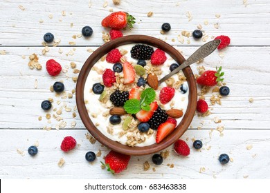 bowl of oat granola with yogurt, fresh raspberries, blueberries, strawberries, blackberries and nuts with spoon on white wooden board for healthy breakfast, top view - Powered by Shutterstock