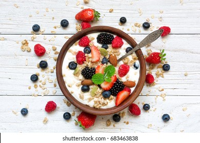 Bowl Of Oat Granola With Yogurt, Fresh Raspberries, Blueberries, Strawberries, Blackberries And Nuts With Spoon On White Wooden Board For Healthy Breakfast, Top View