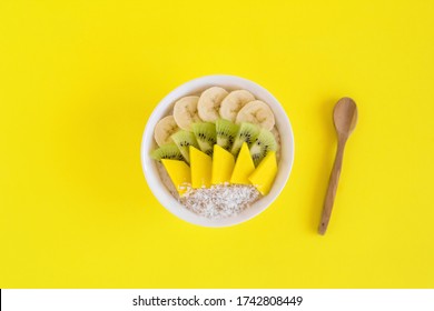 Bowl Of Oatmeal Decorated With Banana, Kiwi, Mango And Coconut Flakes And Wooden Spoon On Yellow Background. Flat Lay With Copy Space.