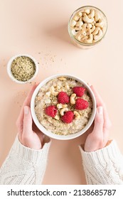 Bowl Of Oatmeal Porridge With Raspberries, Hemp Seeds And Nuts In Female Hands Over Beige Background. Top View. Concept Of Clean Eating, Dieting, Healthy Breakfast Food